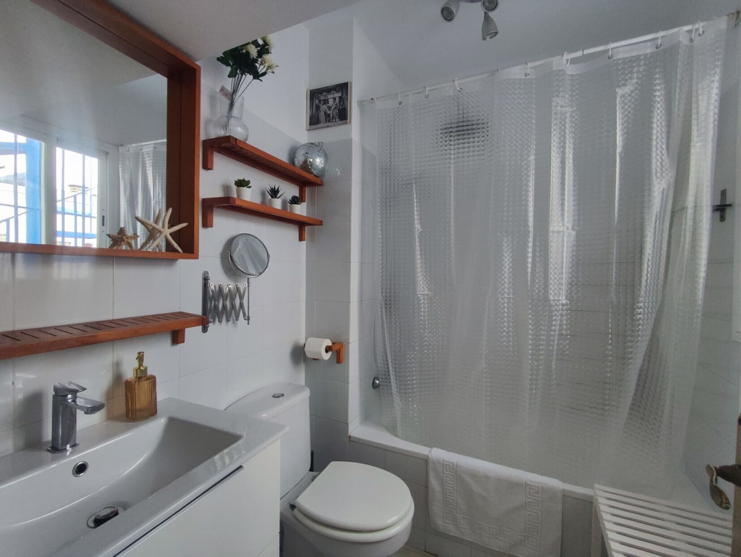 Ein Badezimmer mit weißem Waschbecken, Toilette und Duschvorhang. Holzregale mit Dekorationselementen und ein Spiegel über dem Waschbecken.