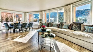 A spacious living room with a large sofa, a dining area with chairs, and a view of a snowy mountain landscape through large windows. - Chalet D'Espoir - Pure Alpine Luxury! (Montriond)