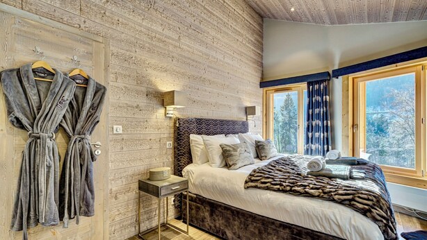 A bedroom with a wooden wall, a bed with a fur throw, and a window with a view of trees. - Chalet D'Espoir - Pure Alpine Luxury! (Montriond)