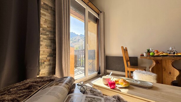 Una habitación acogedora con un ventanal grande, una mesa de madera con cosas para el desayuno, una alfombra mullida y un libro abierto.