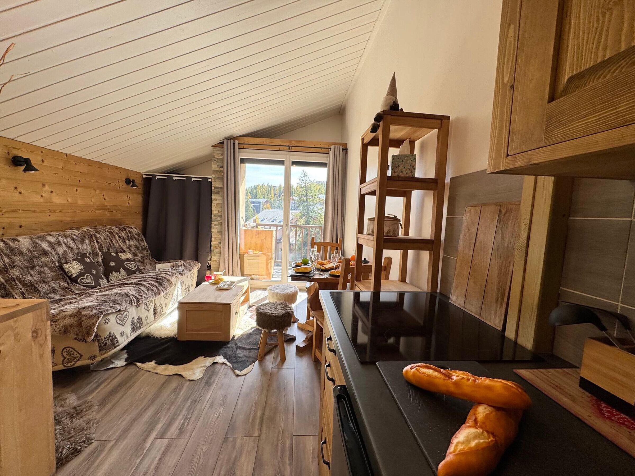 Un accogliente interno di una baita con soffitto in legno, un divano, un tavolo da pranzo con sedie e un angolo cottura con fornello e pane sul bancone.