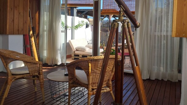 Una terraza de madera con mobiliario de mimbre, un telescopio y vistas a un jardín y un edificio al fondo.