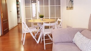 Una habitación con una mesa redonda de madera, sillas blancas, un sofá gris y un ventanal con persianas.