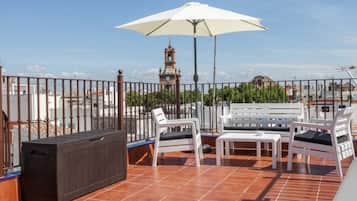 Een terras op het dak met een witte parasol, witte lounge fauteuils en een zwarte terrasverwarmer.