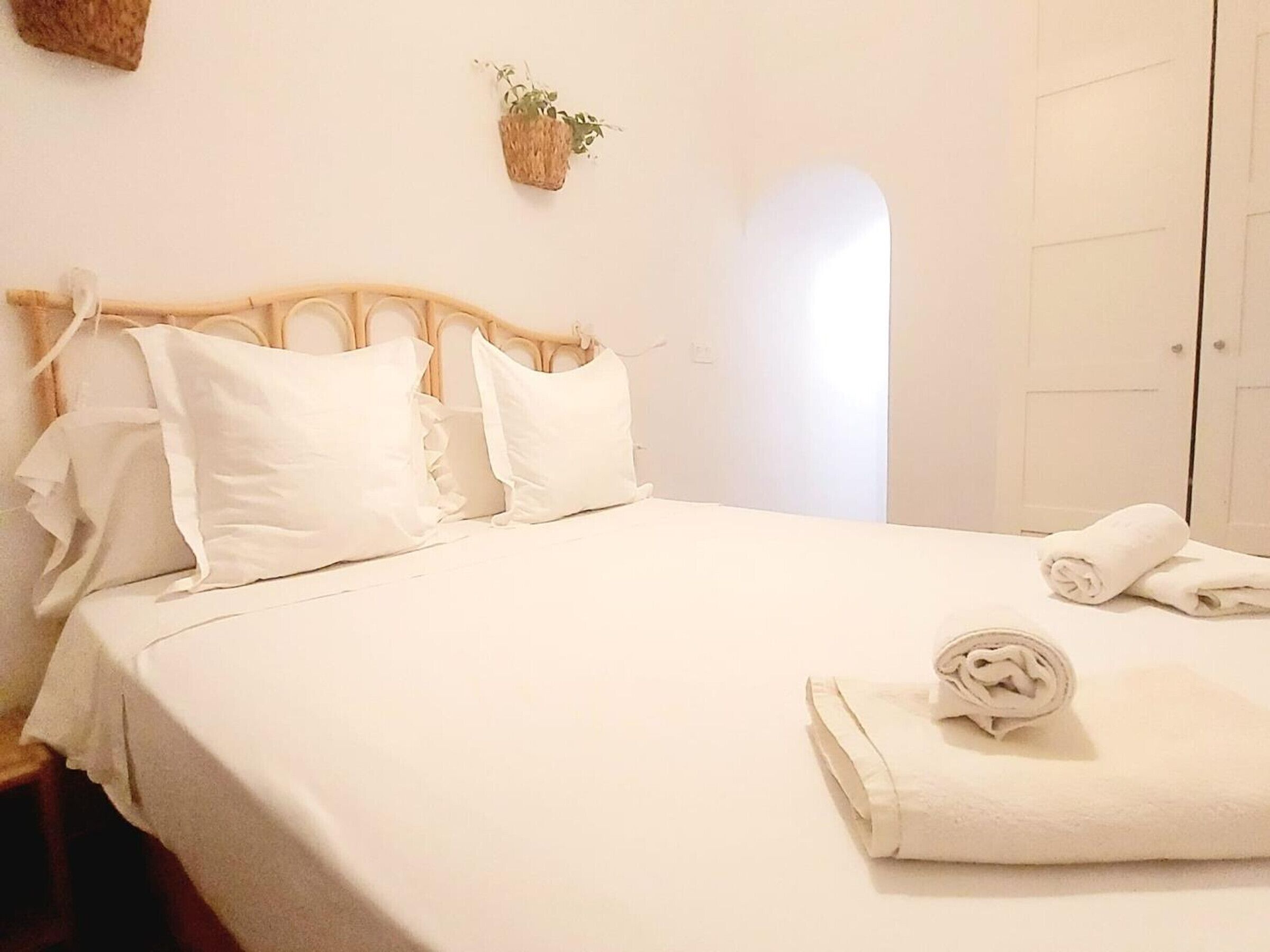 Een netjes opgemaakt bed met witte lakens en kussens, een opgerolde handdoek op het bed, en een kleine plant aan de muur.