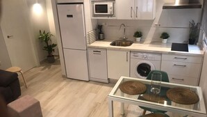 Cocina compacta con refrigerador, microondas, lavamanos y lavadora. Hay una mesa de vidrio con sillas y un pequeño sofá.