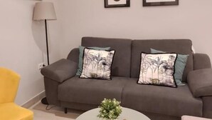 Una sala de estar con un sofá gris, dos cojines estampados, un sillón amarillo y una mesa de centro redonda con una planta.