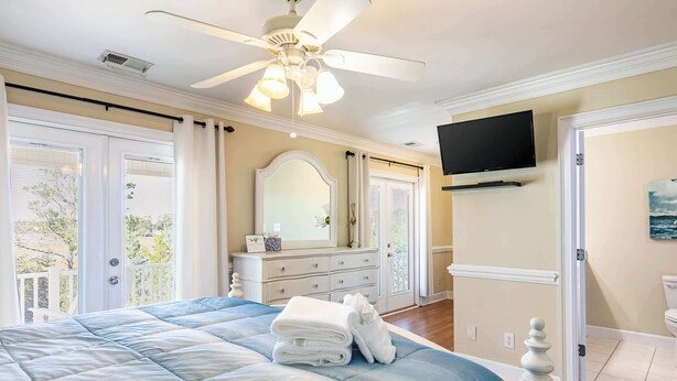 Ein Schlafzimmer mit einem großen Bett, einem Deckenventilator, einer Kommode, einem an der Wand montierten Fernseher und Blick ins Freie durch das Fenster.