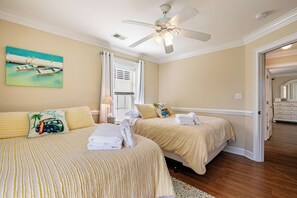 A hotel room with two beds, a ceiling fan, a painting of boats, and a view of the ocean through the window.