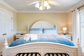 A bedroom with a white bed, blue bedding, a ceiling fan, and a nightstand with a lamp.