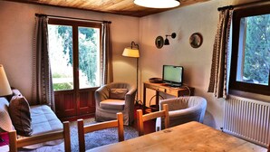 A cozy room with a wooden ceiling, a dining table, a sofa, a chair, a lamp, a computer on a small table, and two windows with curtains. - 6-person apartment in quiet chalet near slopes (Le Grand-Bornand)