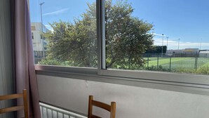 Une pièce avec vue sur un champ verdoyant et des arbres, visible à travers une grande fenêtre. On y trouve une table ronde recouverte d’une nappe et une chaise.