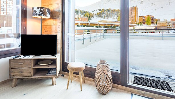 Une chambre moderne dotée d’une grande fenêtre donnant sur un paysage montagneux enneigé, d’un bureau en bois avec un téléviseur à écran plat et d’un petit tabouret.