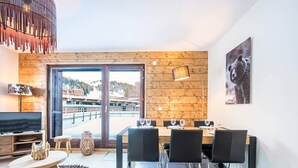 Un espace repas moderne avec une table et des chaises en bois, une grande fenêtre donnant sur des immeubles et une œuvre d’art accrochée au mur.
