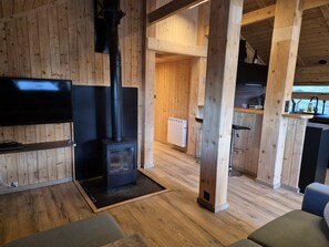 A cozy cabin interior with a wooden interior, a black wood stove, a flat-screen TV, and a kitchen area with a sink and countertop.