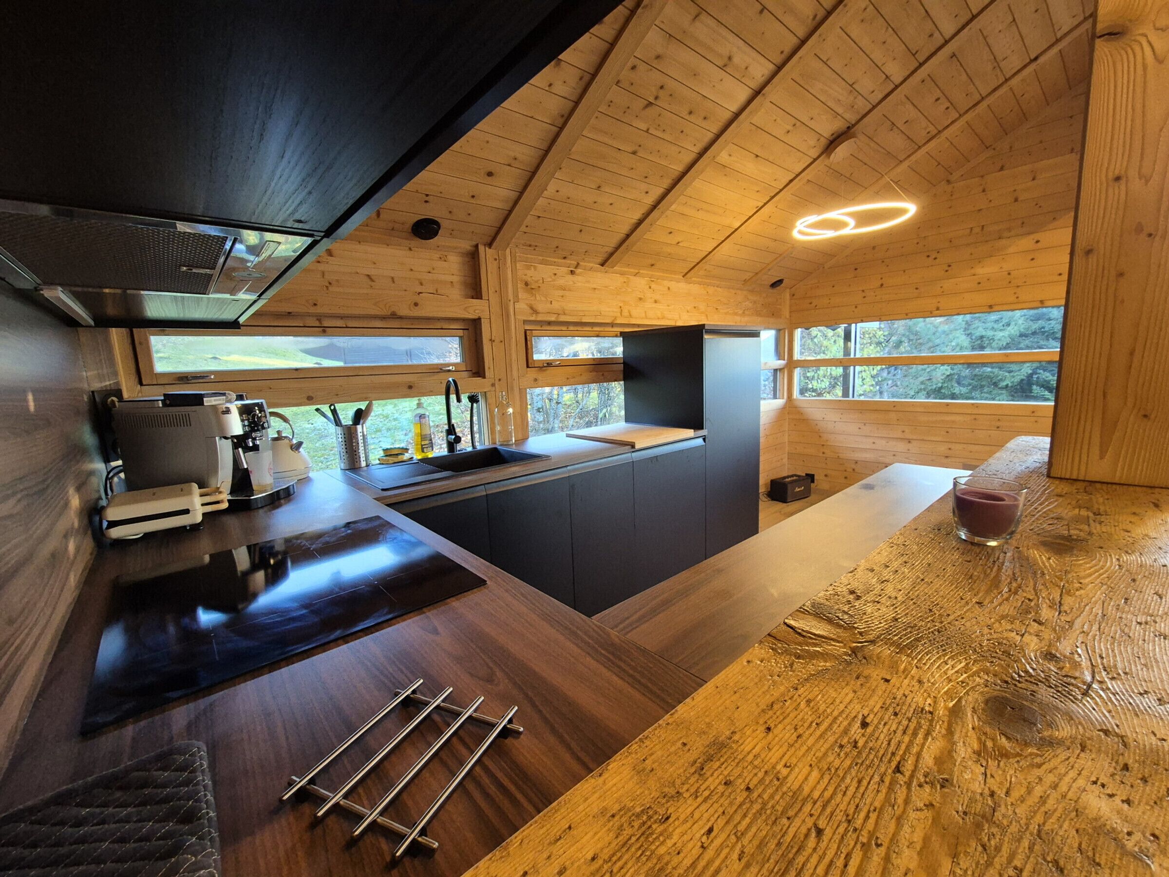 A modern kitchen with wooden beams, a built-in stove, and a large window overlooking greenery.