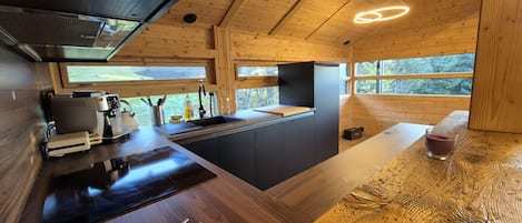 A modern kitchen with wooden beams, a built-in stove, and a large window overlooking greenery.