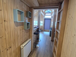 Un couloir étroit aux murs en bois et muni d’un radiateur. On y trouve deux tabourets de bar et une petite table sur laquelle sont posés une bouteille et des verres.