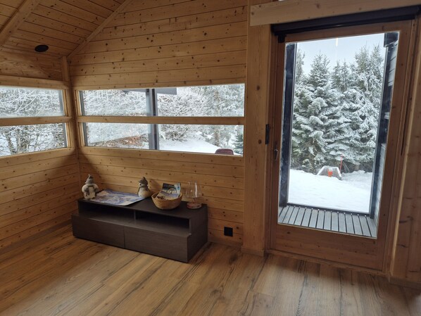 Interior de cabana de madeira com um banco, um cesto e uma janela com vista para uma floresta coberta de neve.