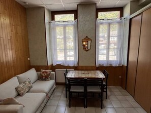 A room with a sofa, a dining table with chairs, a window with a curtain, and a wall-mounted light fixture.