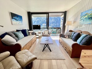 Un soggiorno moderno con divano, poltrona e tavolino. C'è una TV e un balcone con vista sugli alberi.