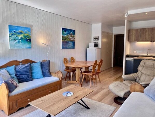 A modern living room with a sofa, a dining table, and chairs. There is a kitchen area with cabinets and a microwave.