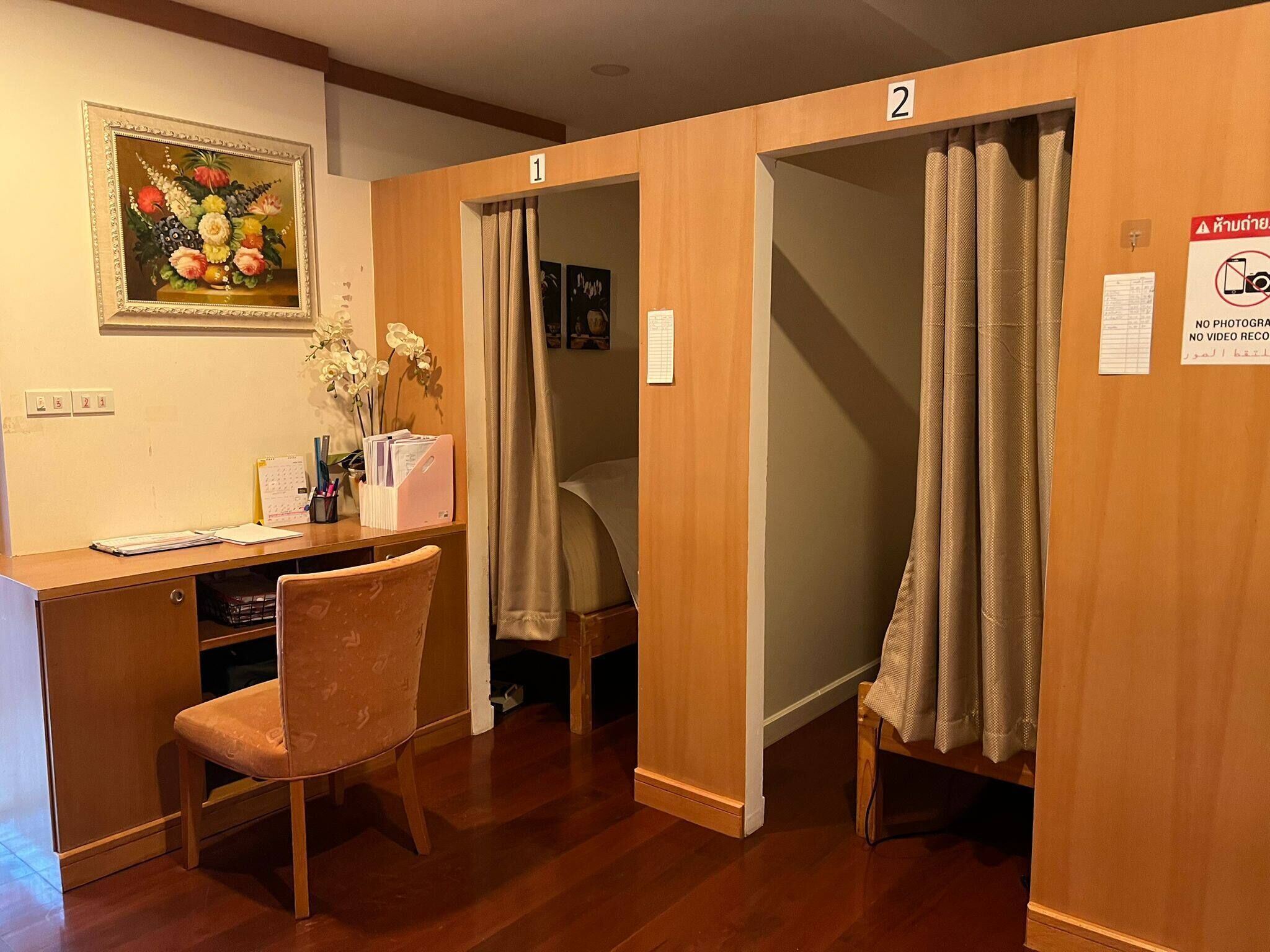 A hotel room with a wooden desk, chair, and a bed with a curtain. There is a painting on the wall and a sign indicating no photography or video recording.
