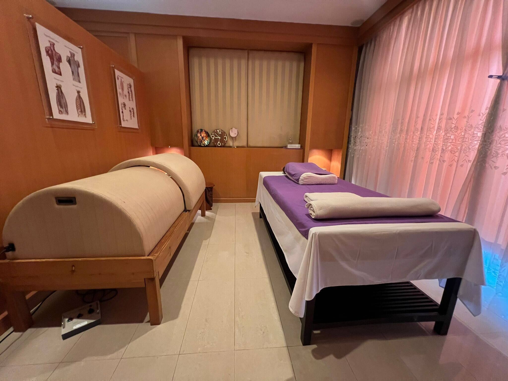 A hotel room with two beds, a window with blinds, a clock, and a wall-mounted shelf with decorative items.