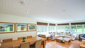 Een ruime kamer met een houten eettafel, rieten stoelen, een beige bank en een groot raam met uitzicht op groen.