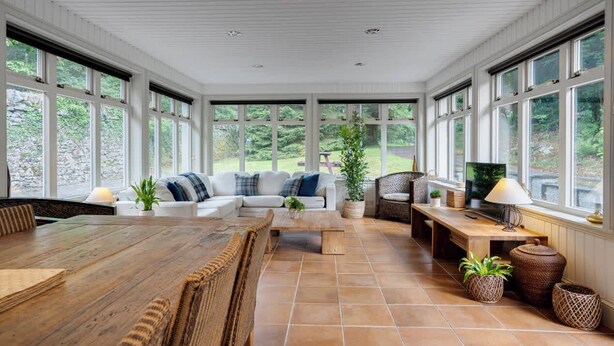 Een zonnehuis met een houten tafel, bank en stoelen, grote ramen en potplanten.