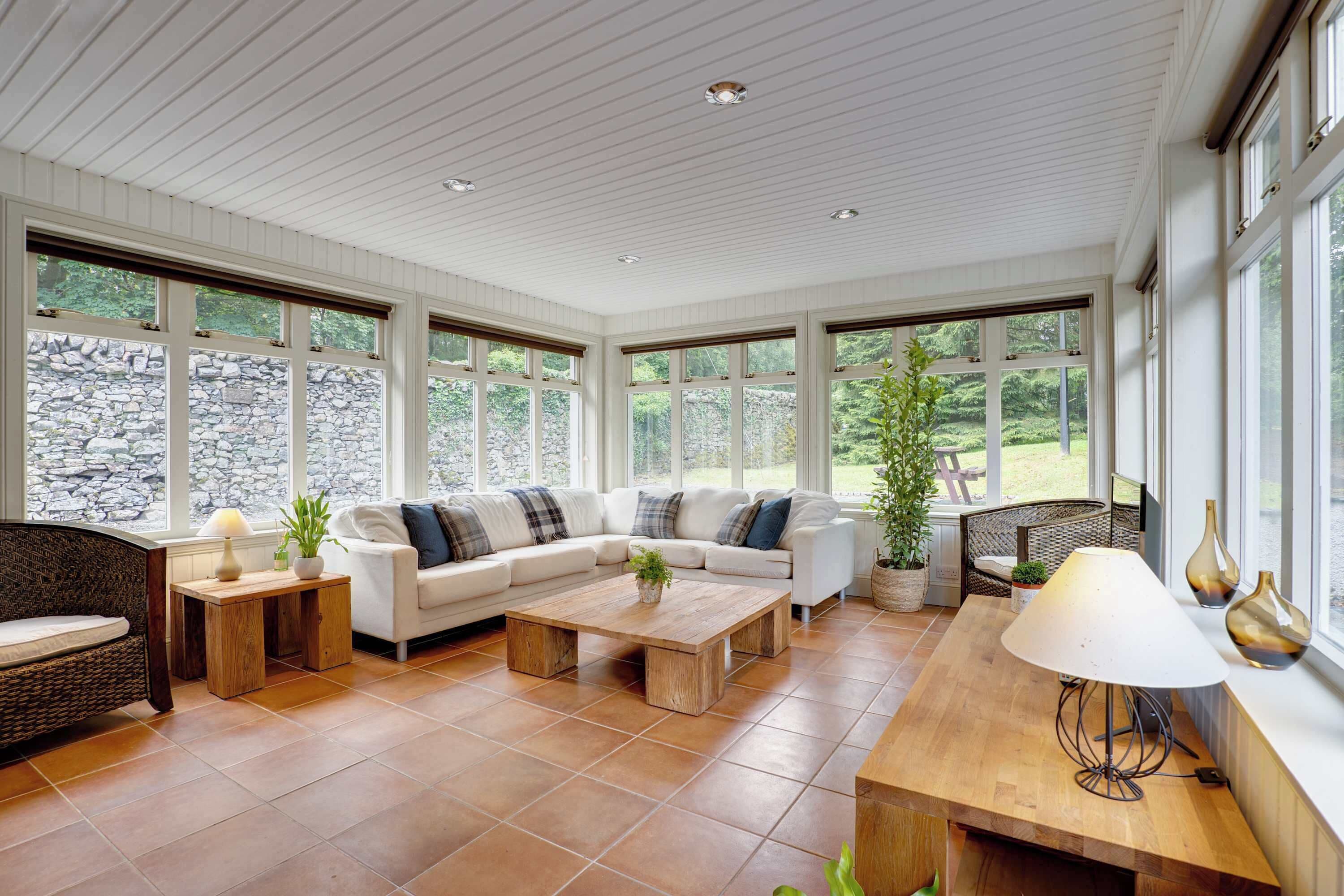A spacious living room with a white sectional sofa, a wooden coffee table, and a dining table with a lamp. Large windows provide views of a stone wall and greenery outside.
