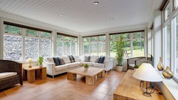 A spacious living room with a white sectional sofa, a wooden coffee table, and a dining table with a lamp. Large windows provide views of a stone wall and greenery outside.