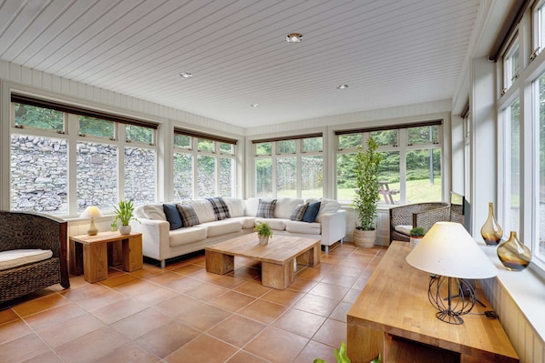 Amplia sala de estar con un sofá modular blanco, una mesa de centro de madera y una mesa de comedor con una lámpara. Ventanas grandes ofrecen vistas de una pared de piedra y vegetación exterior.