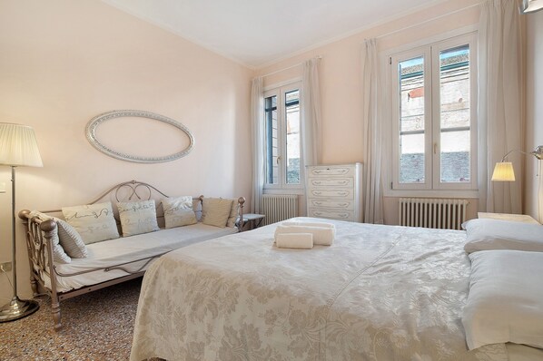 Une chambre à coucher avec un lit, une table de chevet, une lampe, un miroir et une fenêtre avec des rideaux.