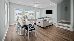 Un salon moderne avec un coin repas, des meubles blancs et une grande fenêtre donnant sur des maisons.
