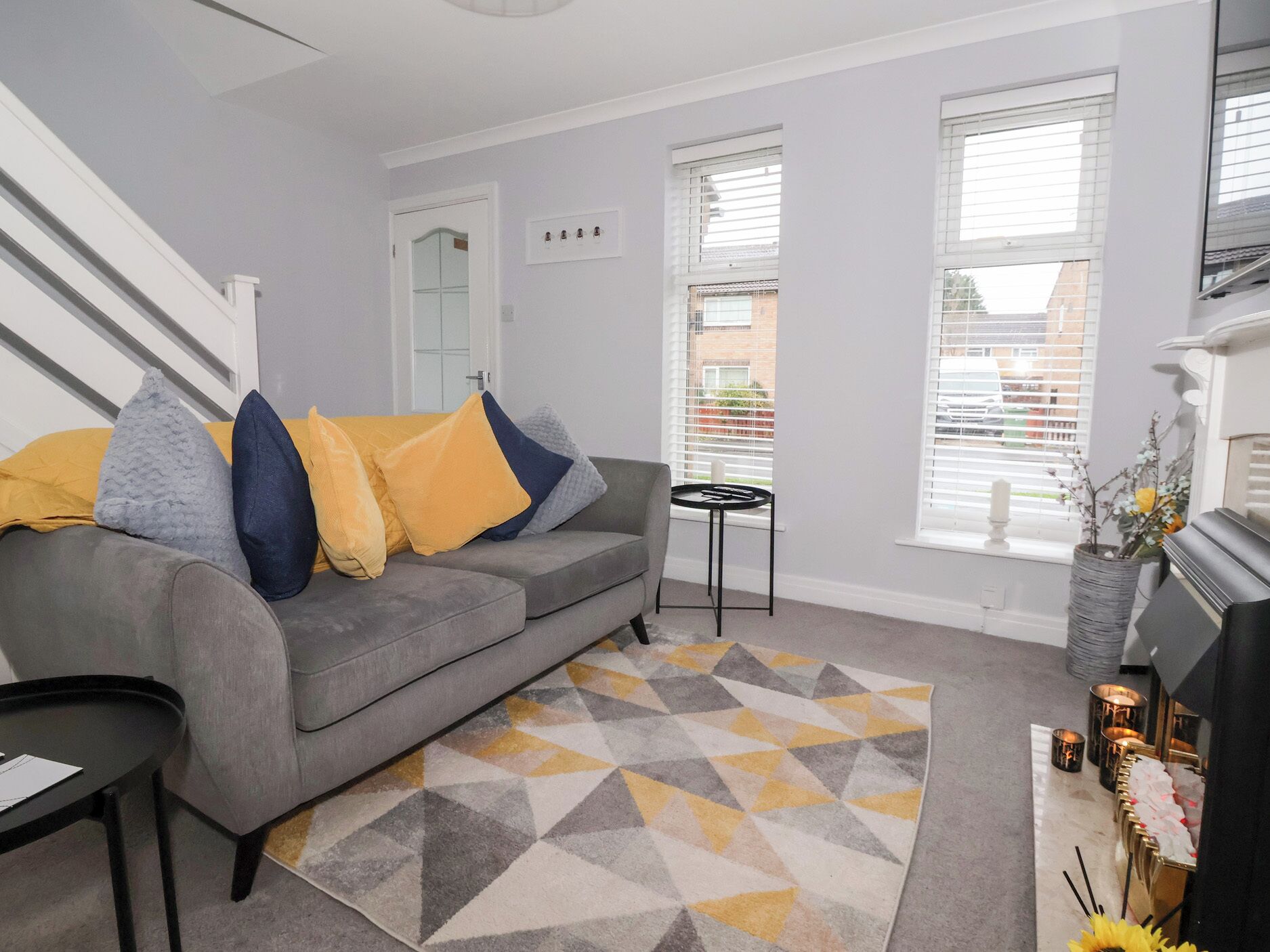 A living room with a grey sofa, yellow and blue cushions, a geometric patterned rug, a small black side table, and a fireplace with candles.