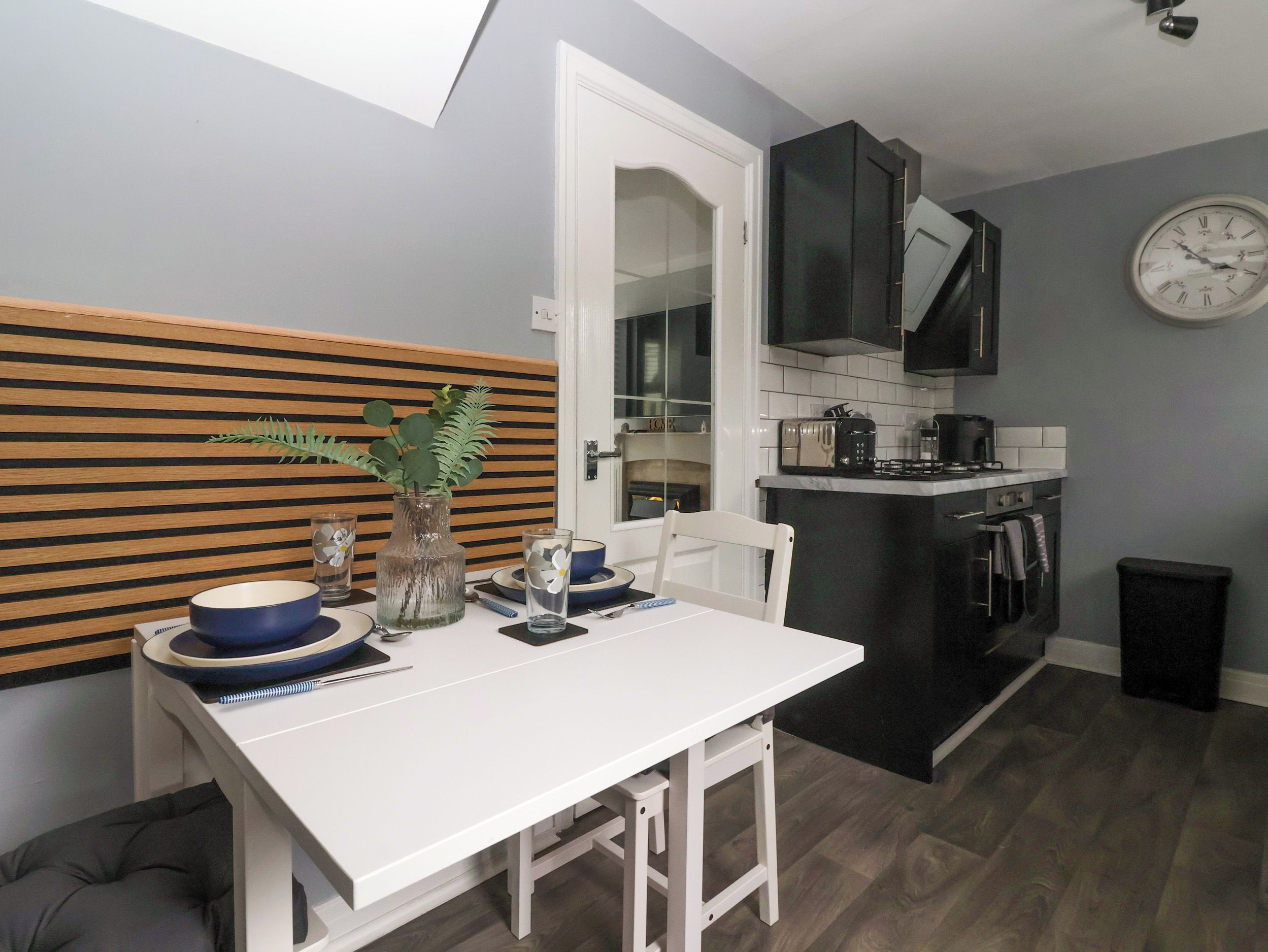 A compact kitchen with a dining area, featuring a white table, a small island with appliances, and a clock on the wall.