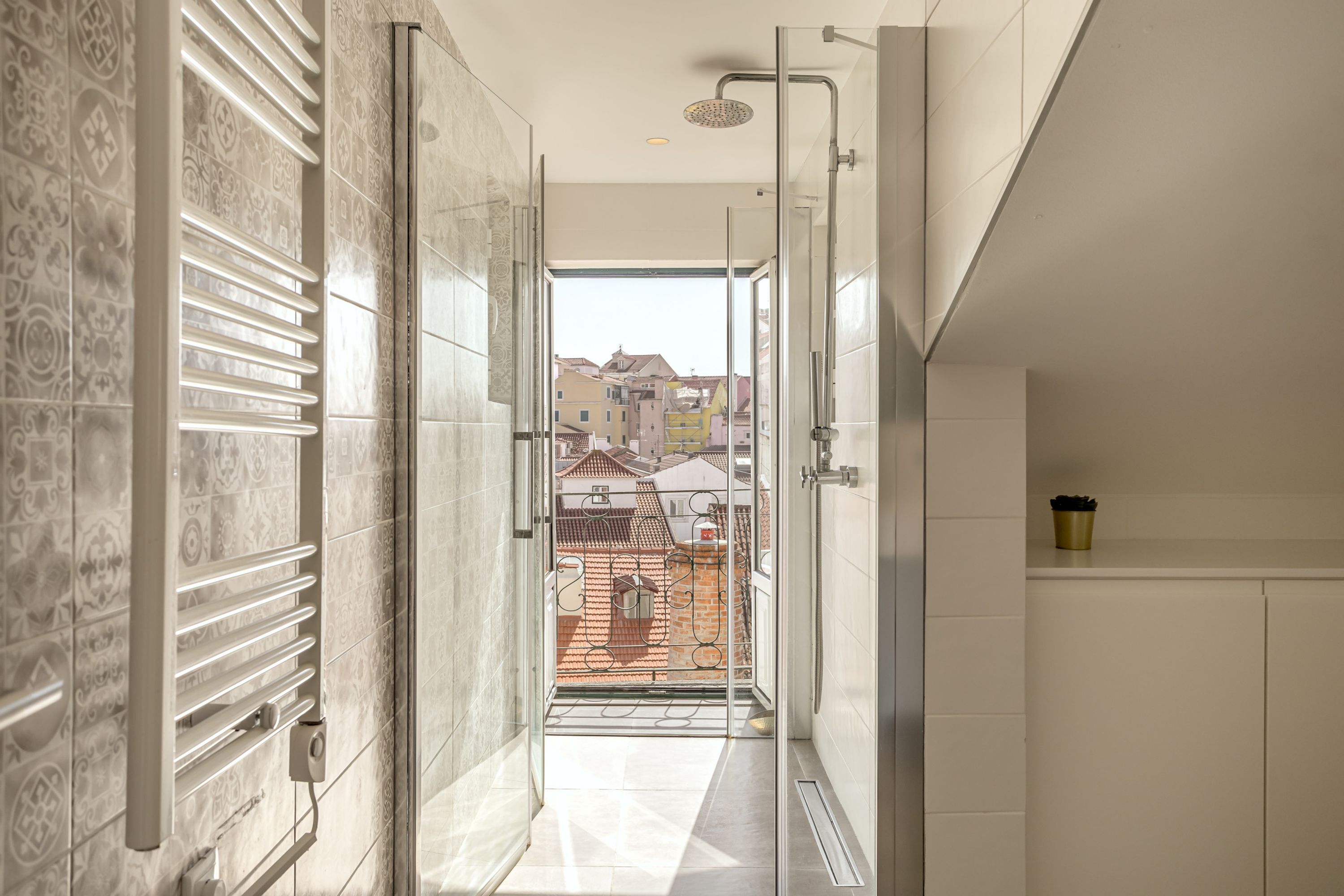 Ein modernes Badezimmer mit einer Glastür, die auf einen Balkon führt, einer Dusche und Blick auf Gebäude.