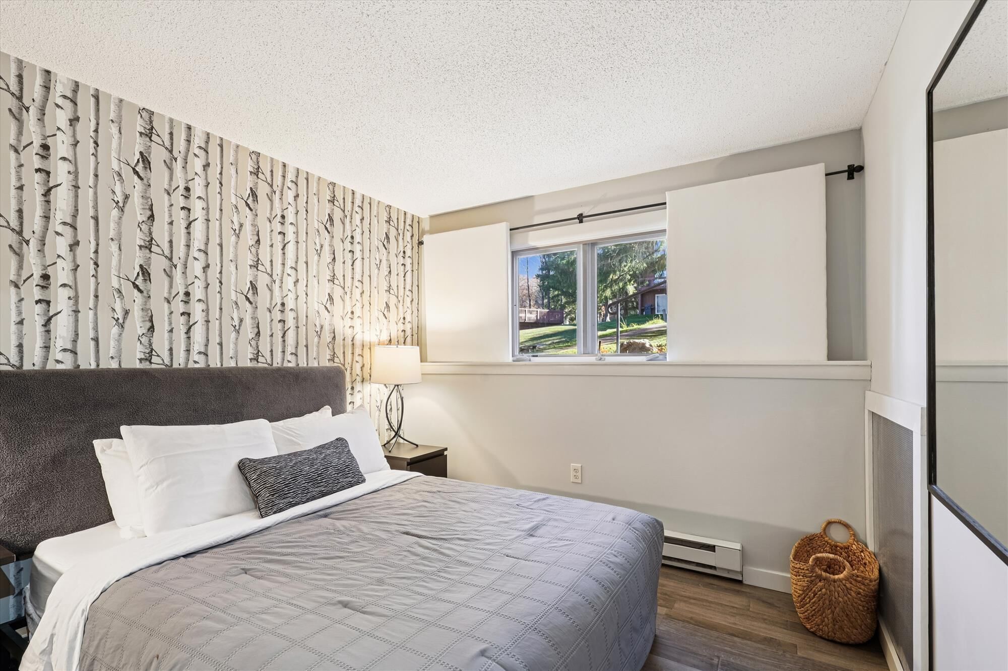 A bedroom with a bed, a window, a wall with a birch tree wallpaper, and a small basket by the window.