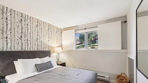 A bedroom with a bed, a window, a wall with a birch tree wallpaper, and a small basket by the window.