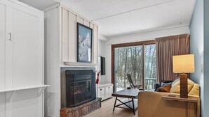 Una sala de estar acogedora con chimenea, un sofá, una mesa y un ventanal con vista a árboles.