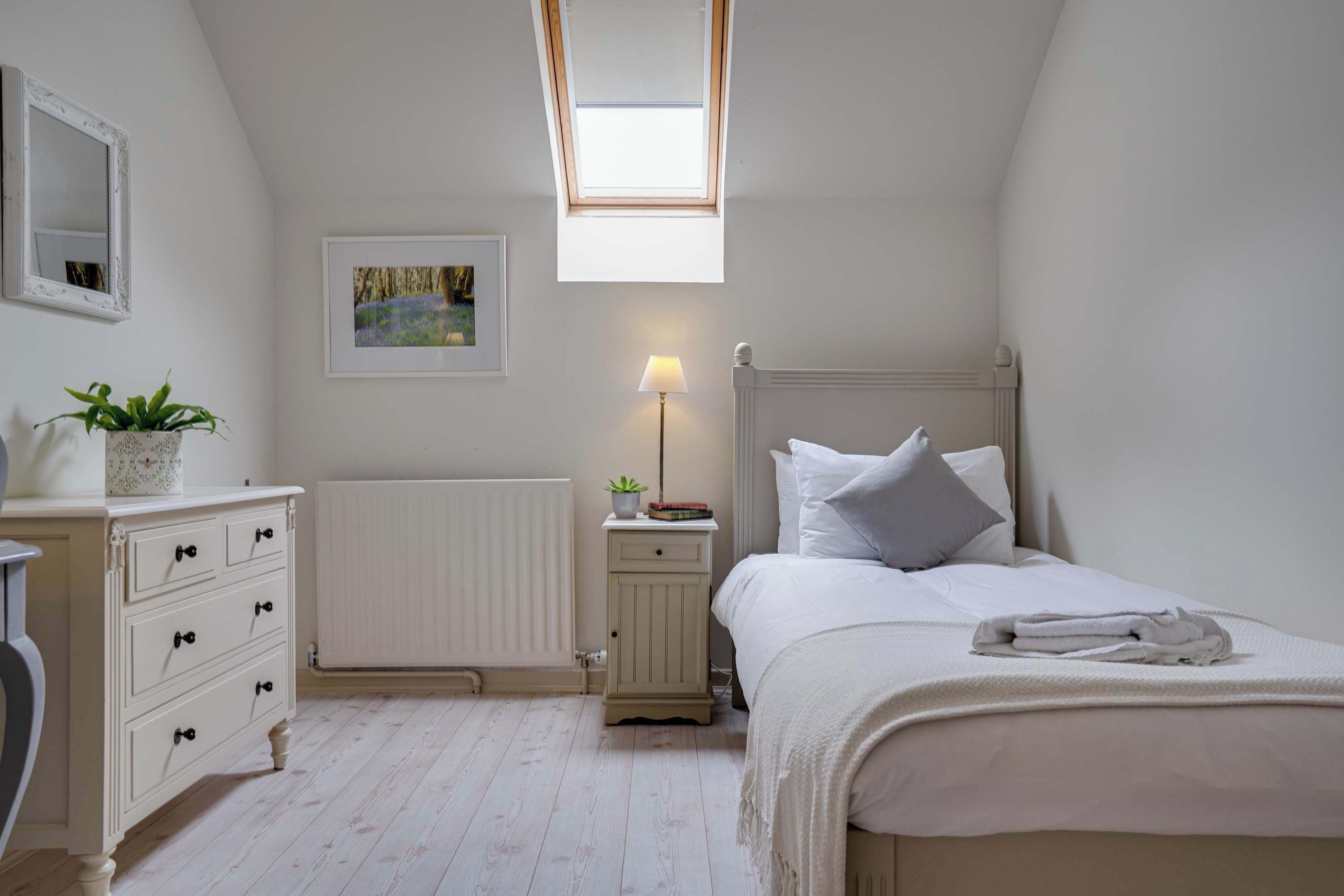A bedroom with a skylight, a bedside table with a lamp, a radiator, and a neatly made bed.