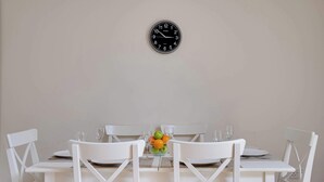 Une table à manger avec des chaises blanches, une horloge au mur et un saladier rempli de pommes et d’oranges.