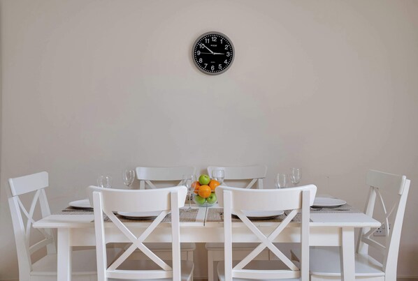 A dining table with white chairs, a clock on the wall, and a fruit bowl with apples and oranges.