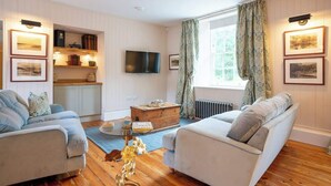 Ein gemütliches Wohnzimmer mit zwei Sofas, einem Couchtisch und einem Fernseher. Ein Bücherregal und ein Fenster mit Vorhängen sind ebenfalls vorhanden.
