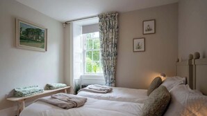 Ein Schlafzimmer mit zwei Betten, einem Fenster mit Vorhängen, einem Heizkörper und einer kleinen Ablage mit gefalteten Handtüchern.