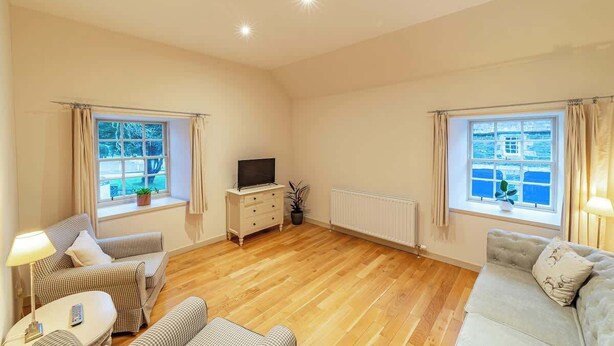 A spacious living room with wooden flooring, two windows with curtains, a television on a dresser, and a sofa. - Penninghame Estate - Bruce Apartment (Penninghame)
