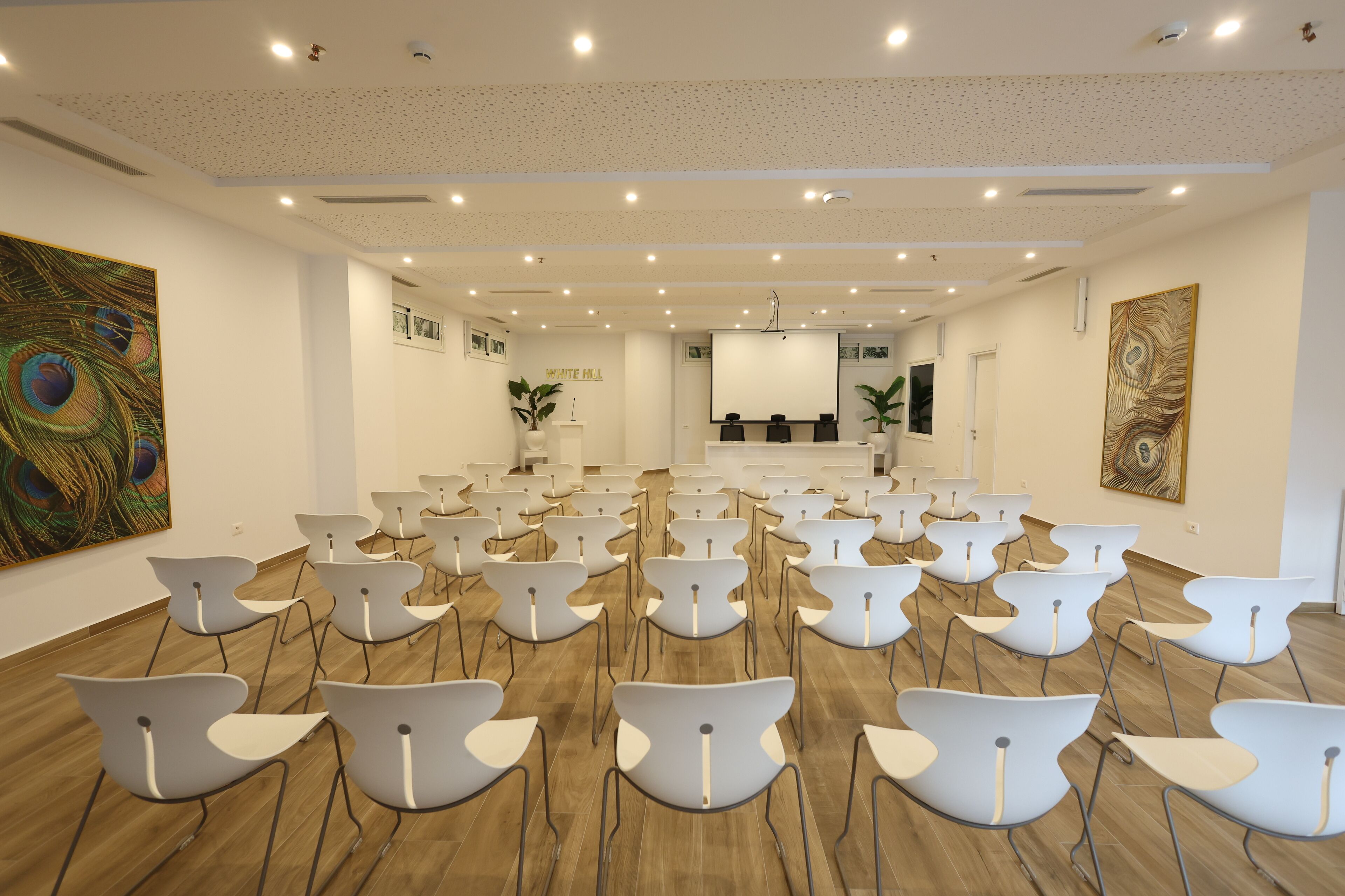 A conference room with rows of white chairs, a stage, and a ceiling with recessed lighting.