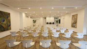 A conference room with rows of white chairs, a stage, and a ceiling with recessed lighting.