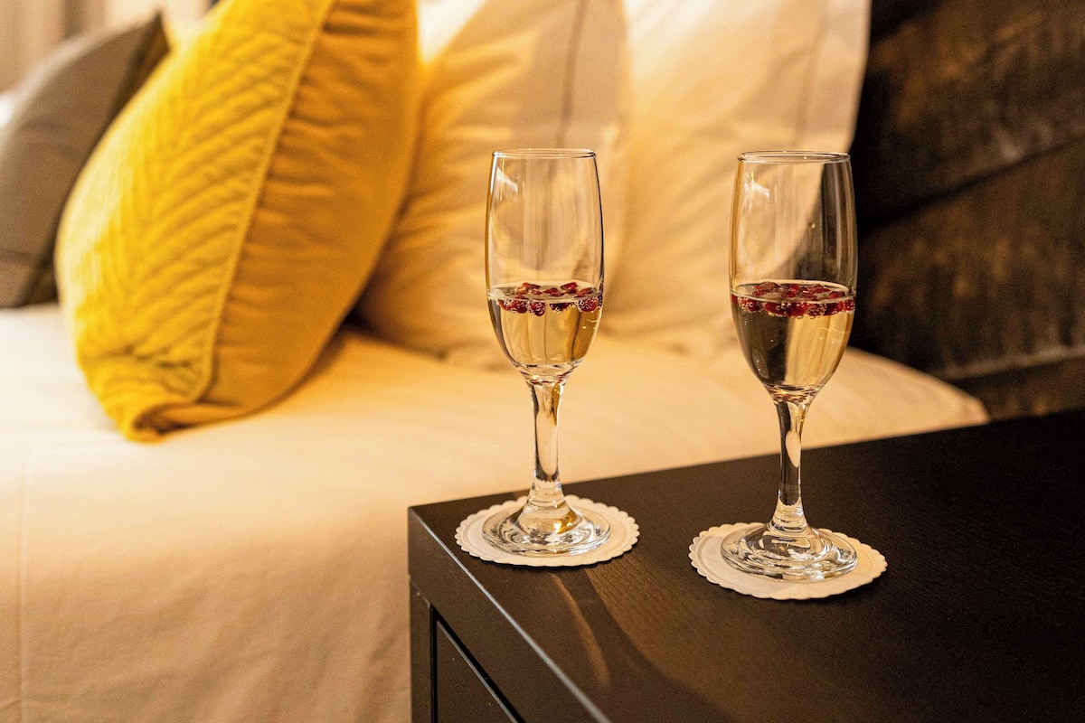A bed with two glasses of wine, a yellow pillow, and a white pillow.